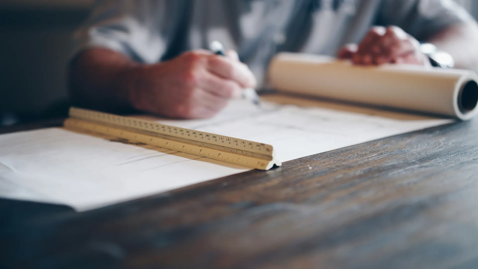 Hands drafting on a drawing with a scale ruler and a roll of plans