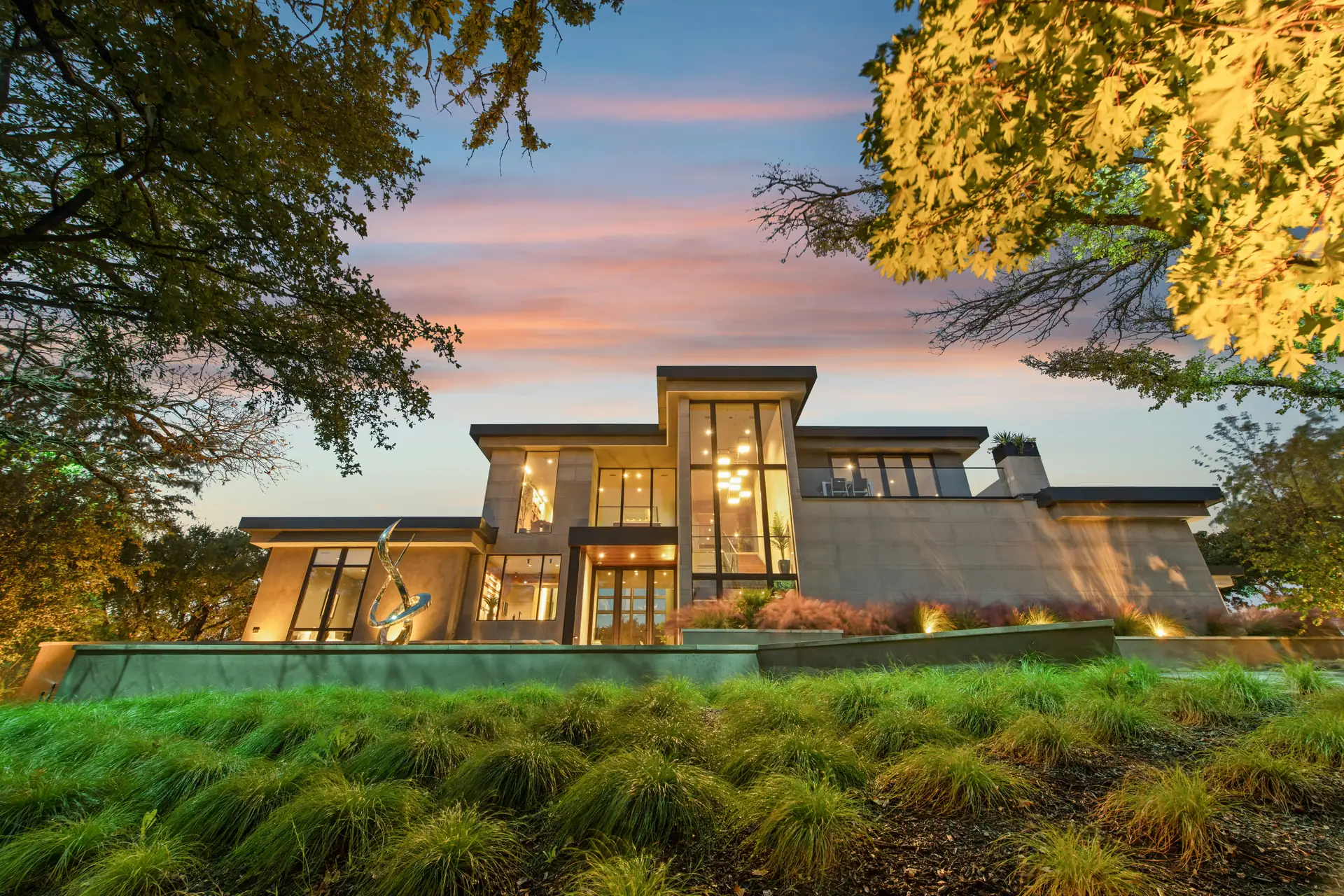 Modern two-story house with lit windows at dusk, behind a bank of ornamental grasses